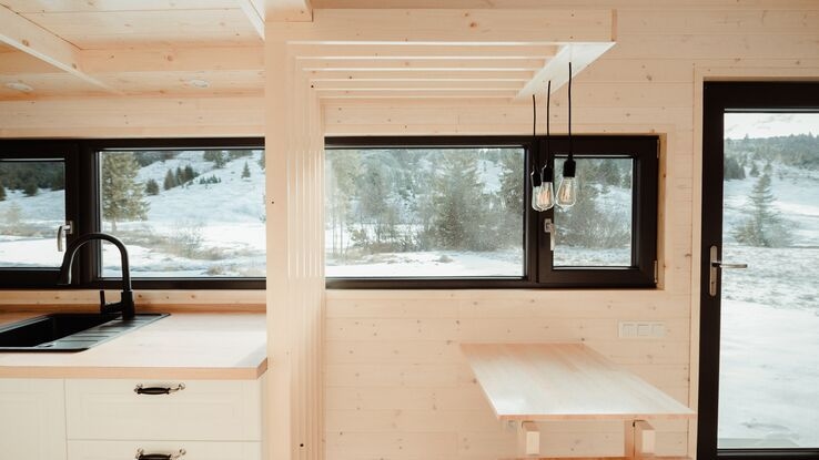 Auf dem Bild ist ein kleines Holzhaus mit großen Fenstern abgebildet. Außerhalb erkennt man eine Wiese, sowie einen Hügel mit Bäumen.
