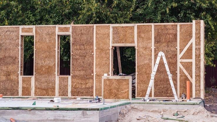 Das Bild zeigt eine Baustelle mit einer teilweise errichteten Wand aus Holzrahmen und Strohballen.