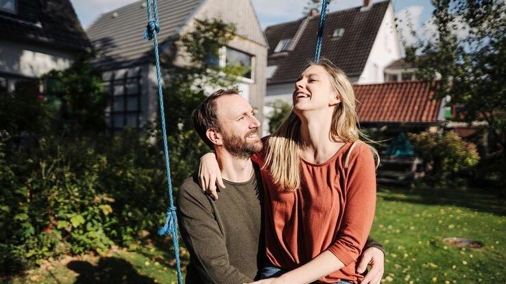 Das Bild zeigt ein Pärchen im Garten auf einer Schaukel. Die Frau sitzt auf dem Schoß des Mannes und beide lächeln sich an.