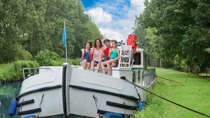 Das Foto zeigt vier Personen auf einem Hausboot. Sie sitzen an der Front des Bootes und umarmen sich. Das Boot ist noch mit Seilen an Pfosten festgemacht.