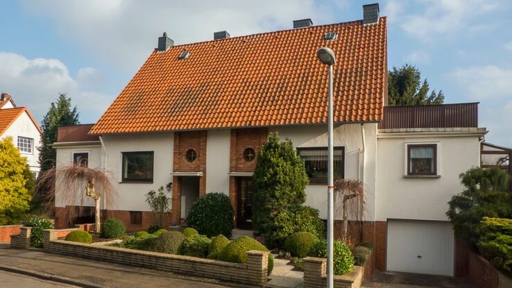 Das Foto zeigt ein älteres Doppelhaus. Vor dem Haus ist ein kleiner Vorgarten mit Pflanzen und einem kleinen Weg zu erkennen. Außerdem ist sowohl links, als auch rechts von dem Haus eine Garage.