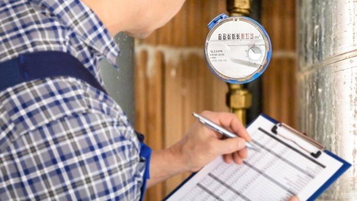 Ein Handwerker erfasst die Wasser- und Stromzählerstände und trägt die Werte mit einem Kugelschreiber in das Formular auf seinem Klemmbrett ein.