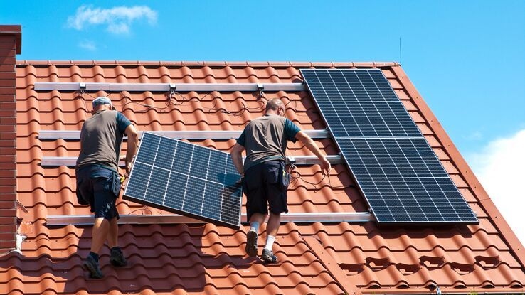 Zwei Handwerker heben gemeinsam ein Solarpanel auf das Dach eines Gebäudes, um es dort zu montieren.