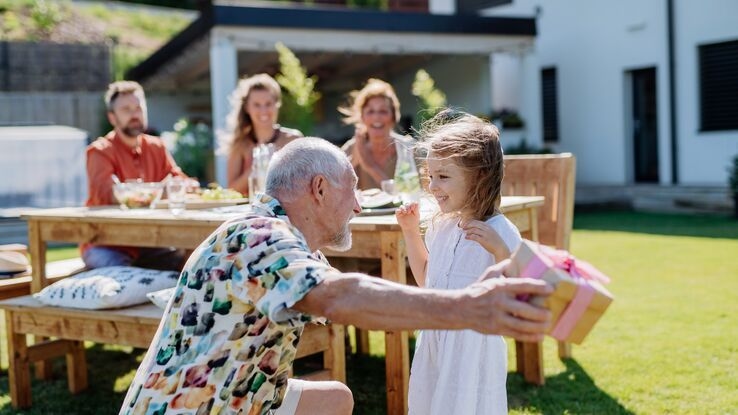 Das Foto zeigt eine Familie in einem Garten. Im Vordergrund übergibt der Großvater seiner Enkelin ein Geschenk. Im Hintergrund sitzen die Eltern des Kindes, sowie die Großmutter an einem Tisch.
