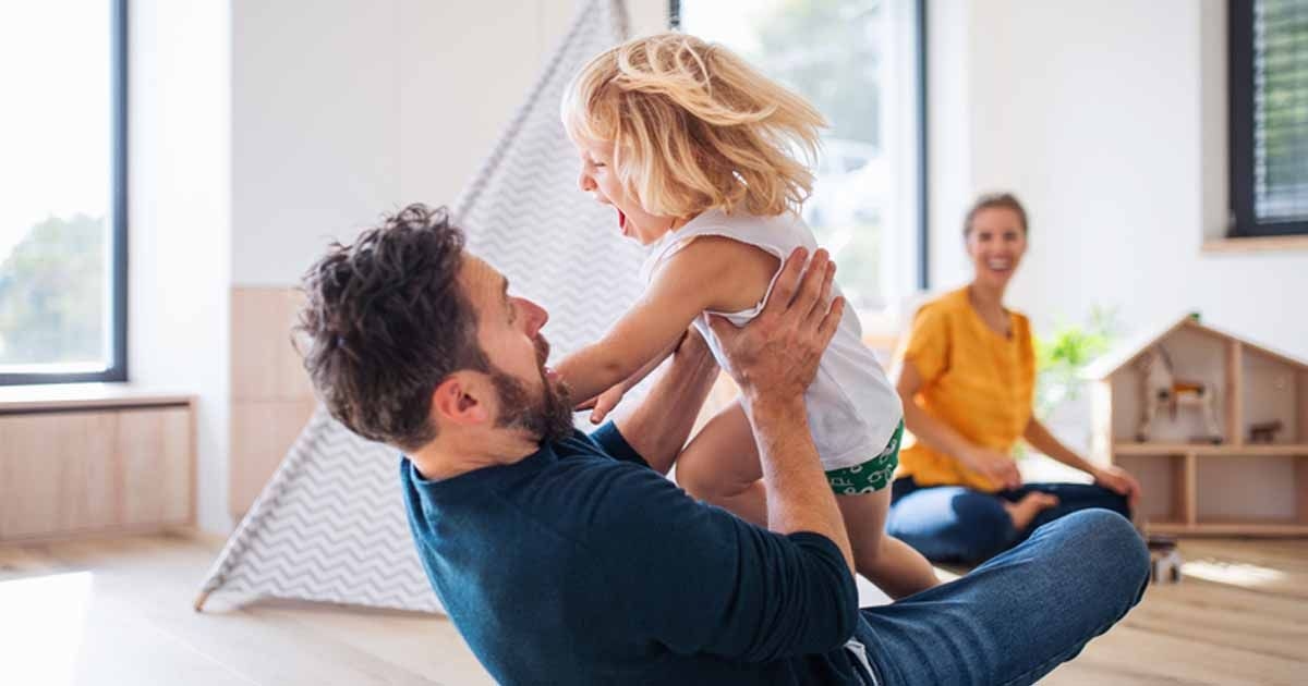 Ein Vater spielt mit seinem Kind im Zimmer. Die Mutter sitzt im Hintergrund und lacht.
