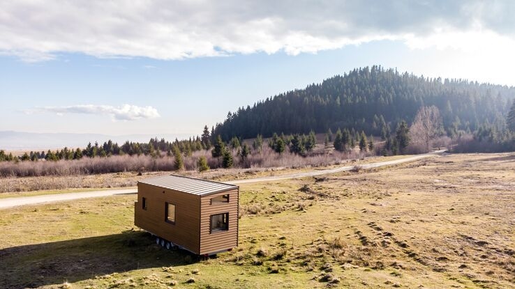 Das Foto zeigt ein Caravan auf einem Feld. Es ist ein kleiner Wohnwagen aus Holz der auf Rädern steht. Im Hintergrund ist ein Hügel mit einem Wald zu sehen.
