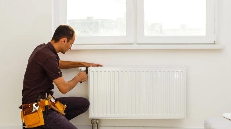 Ein Handwerker kniet an einer Heizung mit einem Werkzeuggürtel um die Hüfte und einem Werkzeug in der Hand. Er ist mit dem Heizungseinbau beschäftigt.