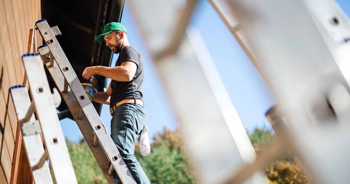 Ein Handwerker steht auf einer Leiter und streicht die Fassade eines Hauses.