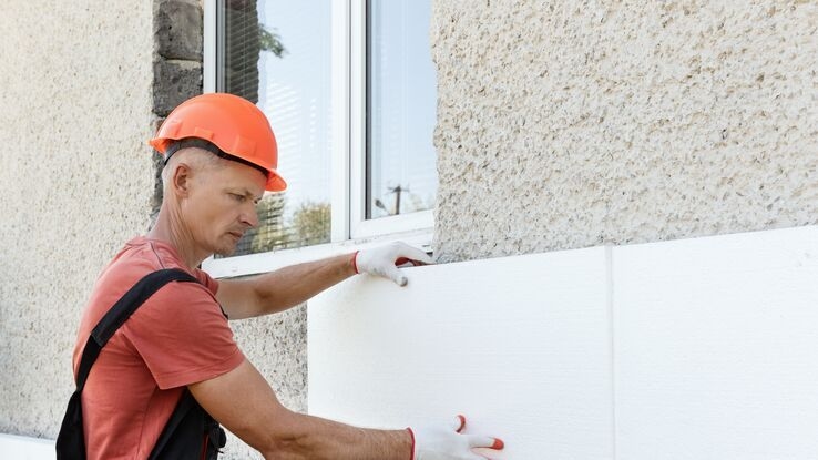 Ein Handwerker modernisiert die Hausdämmung, indem er neue Dämmplatten an der Hauswand anbringt. Er trägt einen Helm Handschuhe und eine Latzhose.