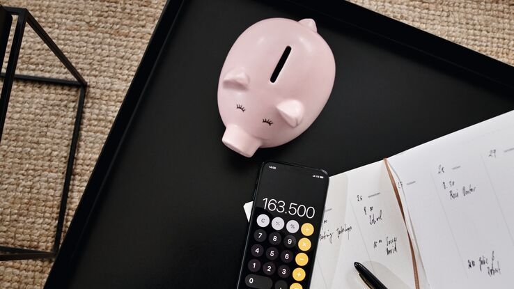 Auf einem schwarzen Tisch liegen ein rosa Sparschwein, ein Smartphone mit geöffneter Rechner-App sowie ein Notizbuch mit handschriftlichen Einträgen. Daneben liegt ein Stift.