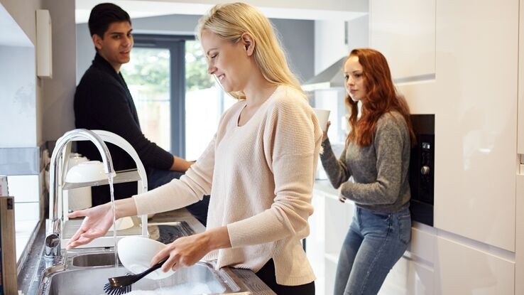 Zwei Frauen und ein Mann stehen in einer Küche. Im Vordergrund spült eine Frau das Geschirr, während die anderen beiden Personen neben ihr stehen.