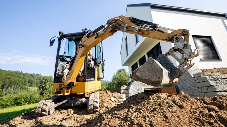 Ein Bagger hebt eine Baugrube aus, dahinter ist ein Neubau zu sehen.
