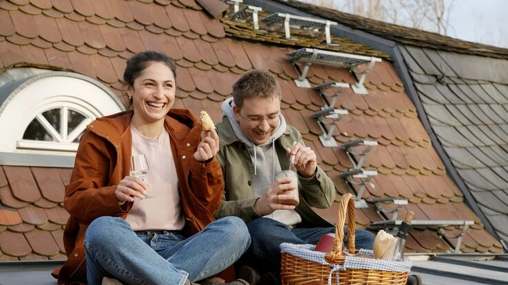 Ein Pärchen sitzt auf einem Hausdach und macht ein Picknick. Sie zelebrieren ihre erste gemeinsame Wohnung.