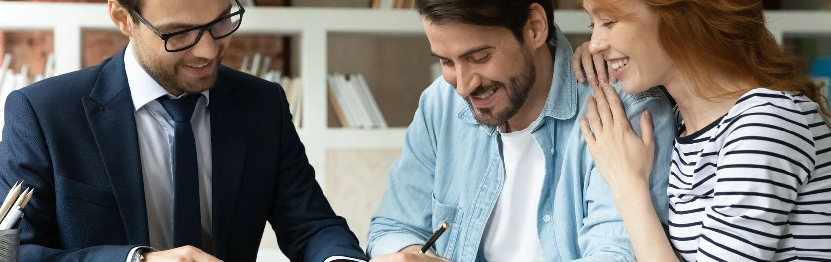 Ein glückliches Pärchen sitzt einem Mann gegebüber und sie schauen auf den Mietvertrag.