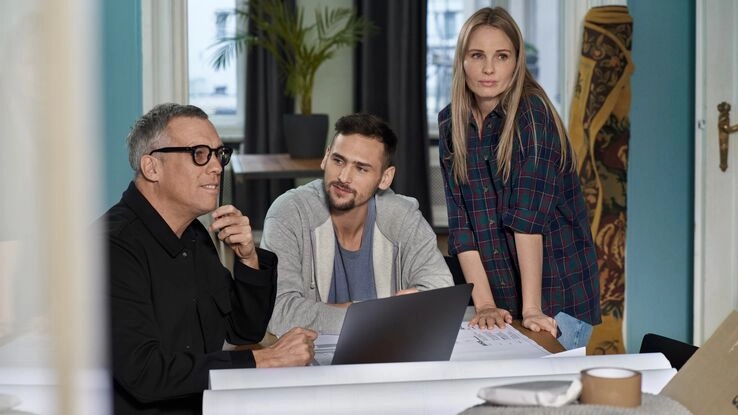 Drei Personen sind um einen Tisch versammelt. Ein Mann mit Brille hat einen Laptop aufgeklappt und der Mann rechts von ihm schaut ihn gespannt an. Die Frau steht neben ihnen und schaut in Richtung des Betrachters. 