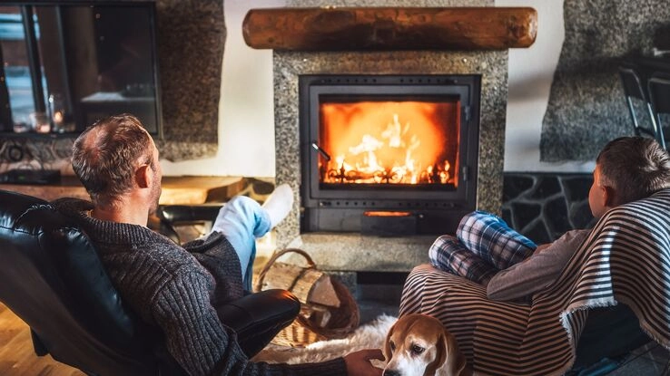 Die Modernisierung des Kamins oder Kachelofens lohnt sich. So steht einem entspannten Abend mit warmer Atmosphäre nichts mehr im Wege, wie man es auf dem Foto sieht. Ein Vater entspannt mit seinem Sohn und Hund vor der Wärme.