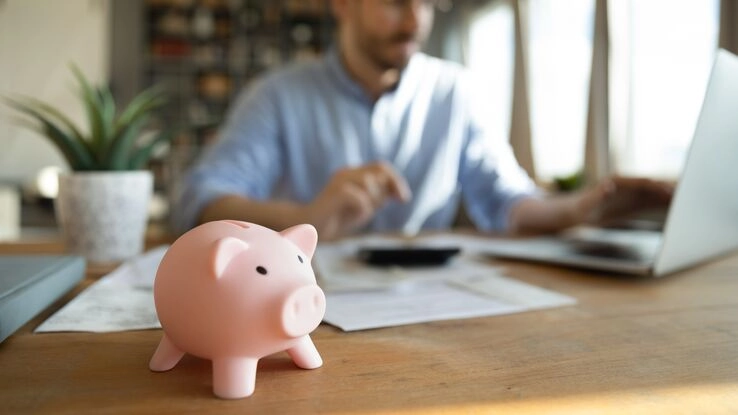 Ein Mann rechnet sich sein Sanierungsvorhaben durch und prüft, wie man Geld sparen kann. Dies symbolisiert ein Sparschwein auf dem Tisch.