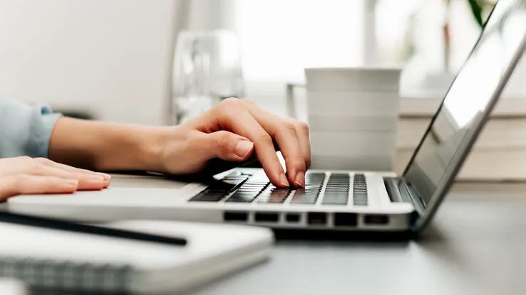 Eine Person tippt auf dem Laptop und hat ein Notizbuch neben sich liegen.