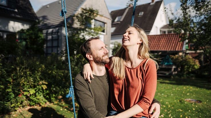Das Bild zeigt ein Pärchen im Garten auf einer Schaukel. Die Frau sitzt auf dem Schoß des Mannes und beide lächeln sich an.