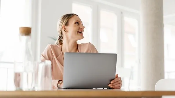 Das Foto zeigt eine junge lächelnde Frau. Sie hat blonde Haare und trägt Ohrringe. Sie sitzt an ihrem Laptop und schaut zur Seite.