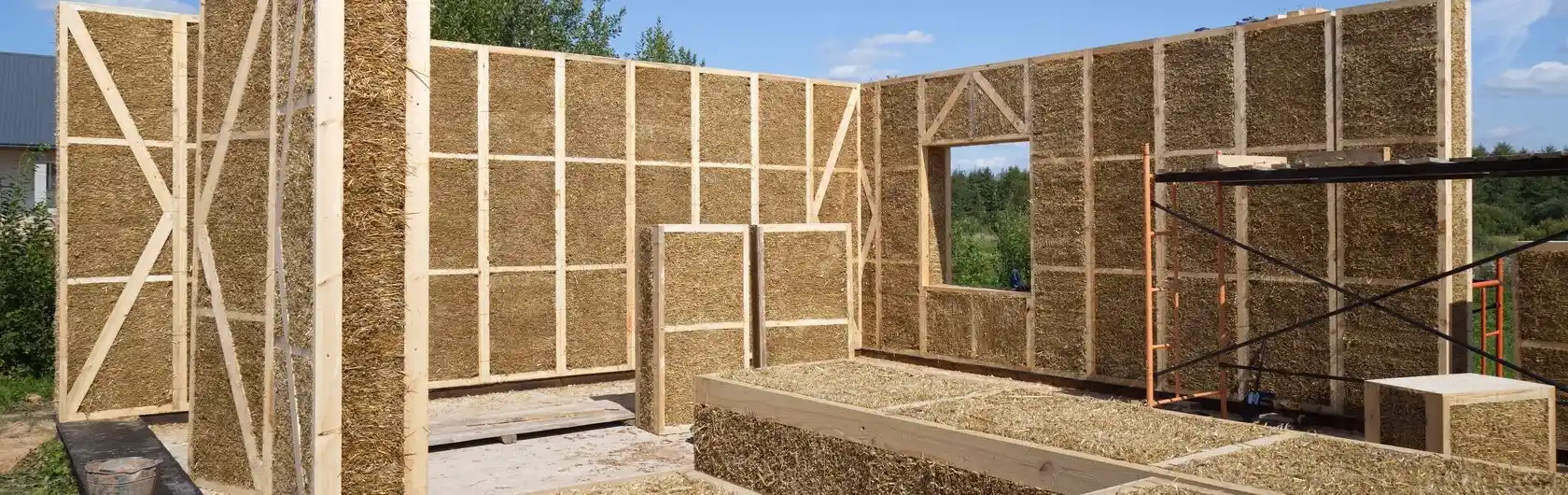 Der Umriss eines umweltfreundlichen Hauses aus Strohballen, bei dem nur die ersten Wände stehen. Das Bild zeigt die Struktur im Bauprozess, umgeben von grüner Natur.