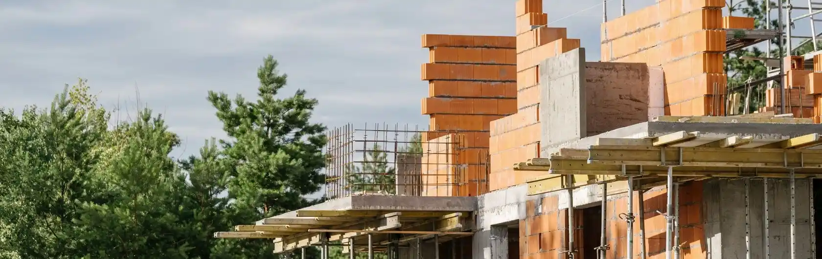 Ein im Bau befindliches Haus, bei dem die Grundstruktur und die ersten Wände bereits stehen. Baumaterialien wie Ziegel und Holz sind um das Gebäude herum verteilt, aber keine Bauarbeiter sind zu sehen.