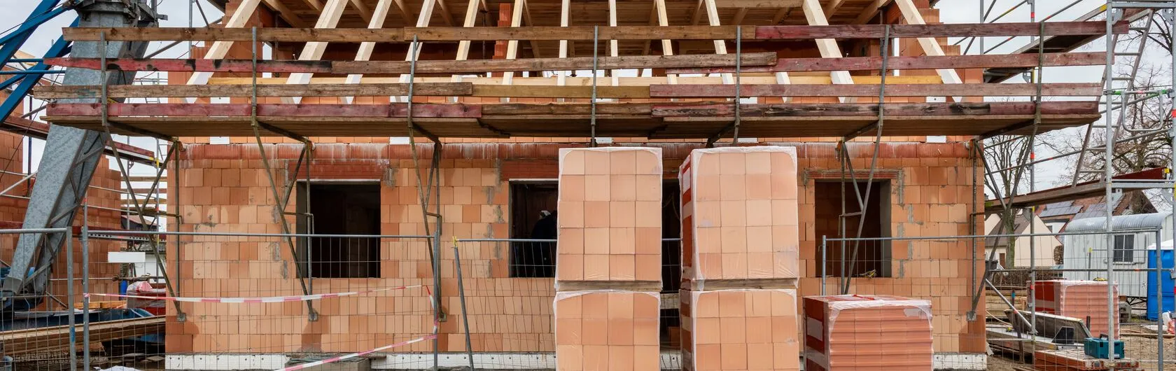 Das Bild zeigt ein im Bau befindliches Haus mit unverputzten Ziegelwänden und einem teilweise errichteten Dachstuhl.