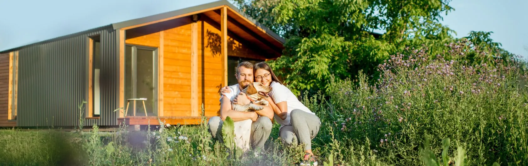 Ein Paar sitzt mit einem Hund vor einem modernen, kleinen Holzhaus, umgeben von üppigem Grün.