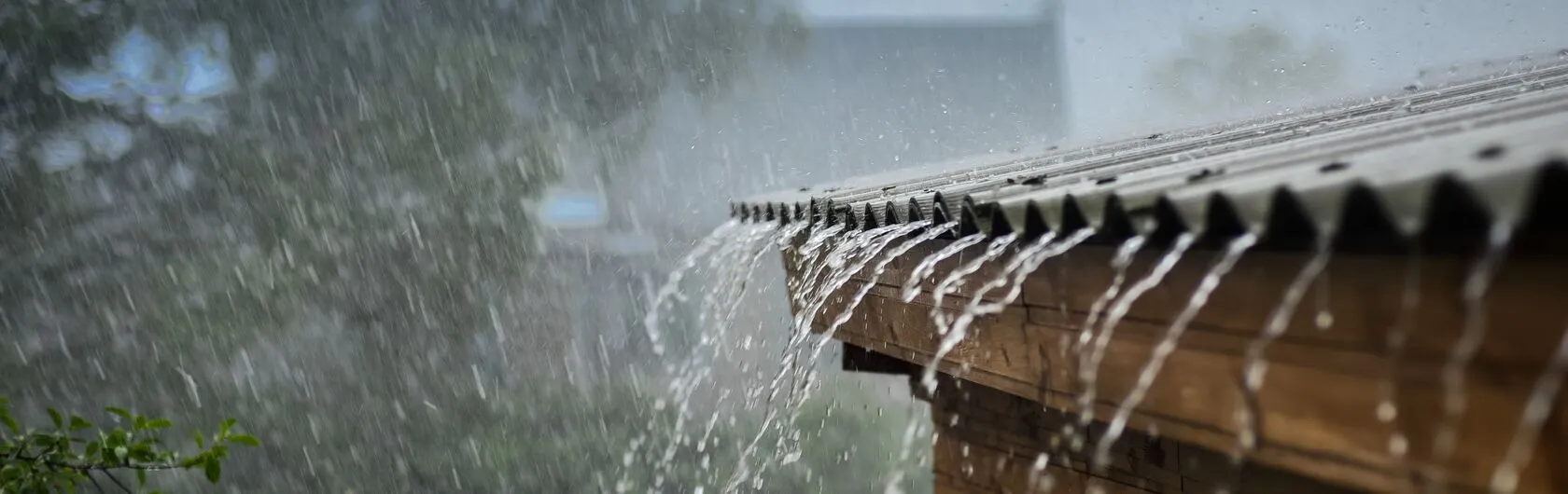 Das Bild zeigt starken Regen, der von einem Wellblechdach auf eine Holzstruktur herabströmt.