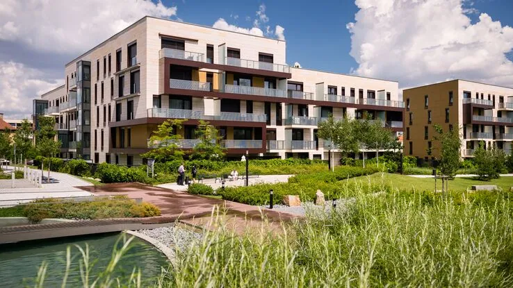 Das Foto zeigt eine moderne Wohnanlage. Es sind einige Wohnkomplexe, sowie ein kleiner Park mit einem Gewässer zu sehen. Die Anlage wirkt freundlich und gepflegt.