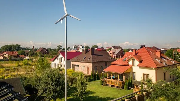 Das Bild zeigt ein kleines Windrad, welches in einem Garten eines Einfamilienhauses steht. Das Wetter ist schön, der Himmel klar und im Hintergrund sind weitere Einfamilienhäuser zu sehen.