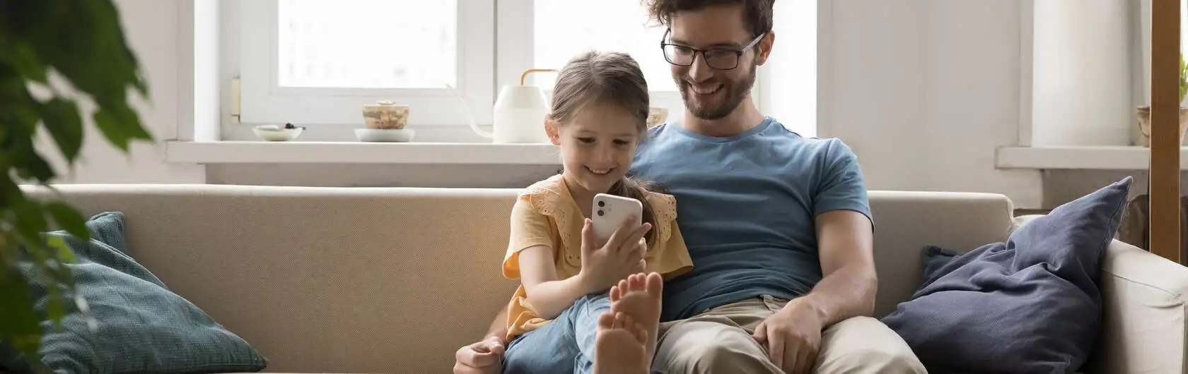 Ein Mann und ein Kind sitzen lächelnd auf dem Sofa und schauen auf das Smartphone, das das Kind in der Hand hält. Beide tragen kurzärmlige T-Shirts. Auf dem Sofa liegen zwei Kissen, und im Hintergrund sind eine Gießkanne und Dekoobjekte auf der Fensterbank zu sehen.