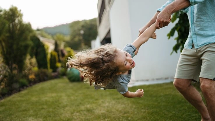 Förderungen bringen Schwung in die Baufinanzierung - ein Vater schwingt sein Kind durch den Garten.