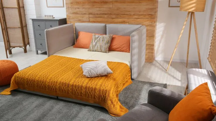Ein Schlafzimmer mit einer gemütlichen Schlafcoach vor einer Holzwand. Auf dem Bett liegen eine orangefarbene Tagesdecke sowie Kissen.