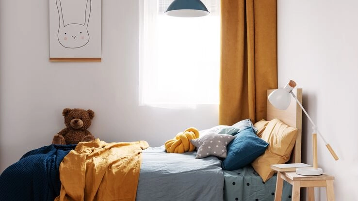 Kinderzimmer mit einem Kinderbett, auf dem viele Kissen liegen und ein Teddybär sitzt.