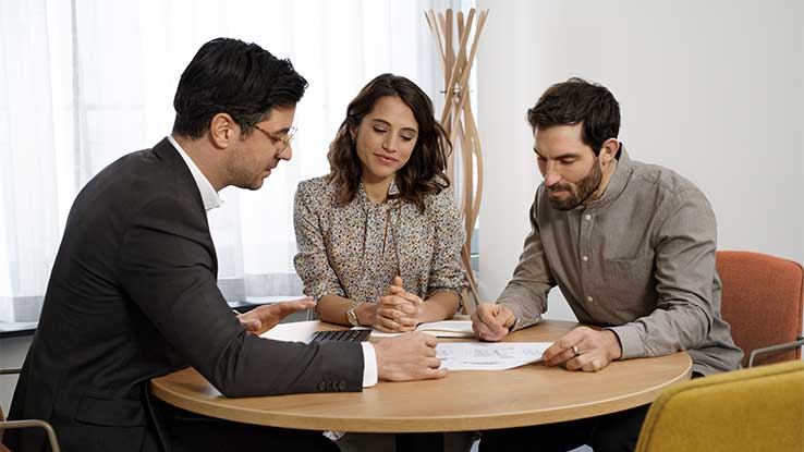 Ein Finanzierungsberater berät ein Pärchen zur Ihrer Immobilien- und Anschlussfinanzierung. Sie sitzen in einem Besprechungszimmer, welches modern ausgestattet ist. Auf dem Tisch liegt ein Finanzierungsplan, auf den sie gemeinsam schauen.