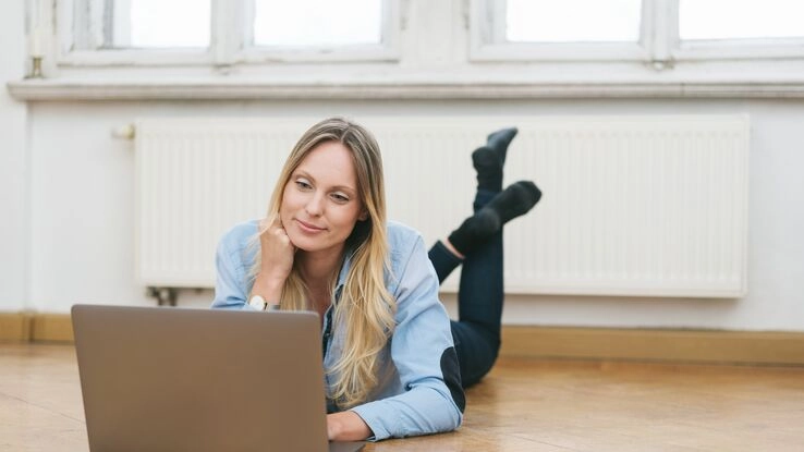 Heizung sanieren? Eine Frau informiert sich über das Thema und liegt dabei an der Heizung vor ihrem Laptop.