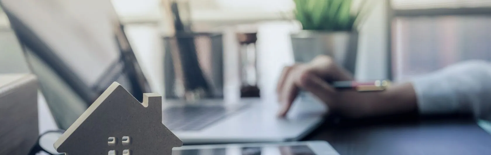 Eine Person sucht im Internet eine Immobilie. Das zeigt ein Laptop sowie ein kleines Haus aus Holz, welches auf dem Schreibtisch steht.