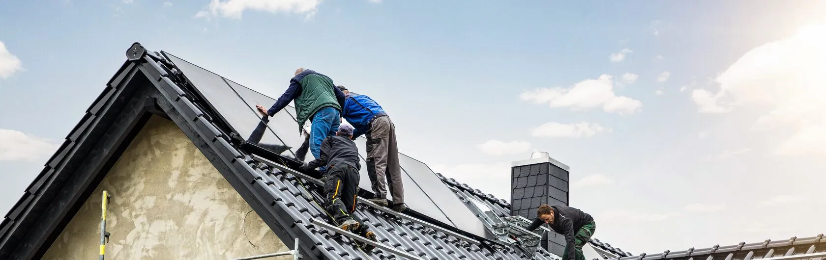 Symbolbild für KfW 296: Bauarbeiter bringen auf dem Dach eine Solaranlage an.